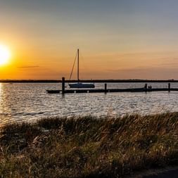 Voilier amarré à une jetée en bois à Rochefort au coucher du soleil. Le soleil doré se reflète sur l'eau calme avec rivage herbeux.