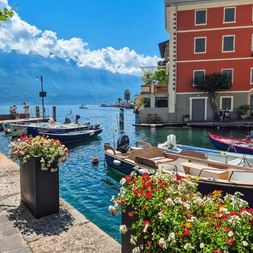 Port pittoresque de Limone avec bateaux colorés amarrés dans l'eau turquoise, jardinières fleuries sur le quai en pierre et bâtiment rouge.