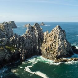 Formations rocheuses spectaculaires à la Pointe de Pen-Hir à Crozon s'avançant dans l'océan turquoise avec vagues blanches et ciel bleu.