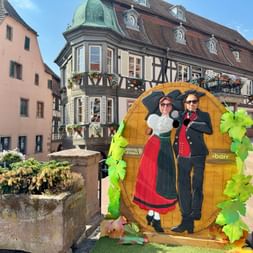 Panneau photo en bois avec couple alsacien en costume traditionnel, décoré de vignes, sur une place avec maisons à colombages.