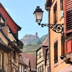 Maisons à colombages traditionnelles sur la Route des Vins d'Alsace avec lampadaire orné et château sur colline avec vignobles en arrière-plan.