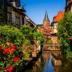 Canal à Wissembourg avec fleurs rouges, maisons à colombages et clocher gothique se reflétant dans l'eau.