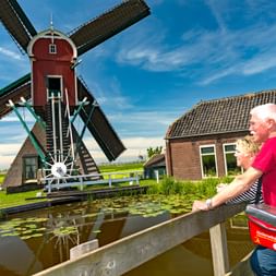 Deux cyclistes avec des sacs observent un moulin hollandais traditionnel rouge et blanc près d'un canal en Hollande du Nord sous un ciel bleu.