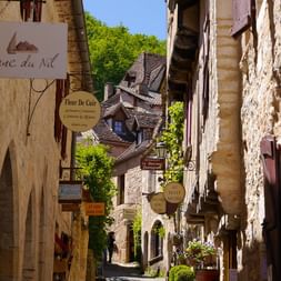 Rue pavée étroite à Saint-Cirq-Lapopie avec bâtiments en pierre, enseignes de boutiques et toits de tuiles traditionnels sur fond de colline.