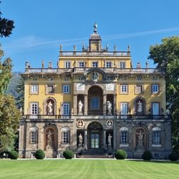 Villa Torrigiani, un palais baroque jaune en Toscane avec des statues sur les balustrades, une entrée voûtée et une pelouse entretenue avec des arbustes ronds.