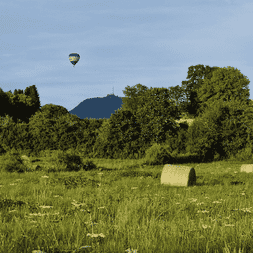 Montgolfière flottant au-dessus d'un pré vert avec des balles de foin. Le Puy de Dôme avec antenne visible en arrière-plan, entouré d'arbres.