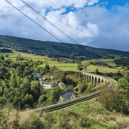 Viaduc ferroviaire en pierre traversant la campagne verdoyante du chemin Stevenson. Collines et forêts sous un ciel bleu avec nuages.