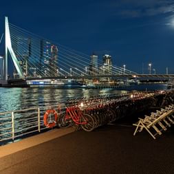 Vue nocturne depuis le pont du MS Normandie montrant l'horizon de Rotterdam avec le pont Erasme illuminé, vélos alignés sur le pont et lune.