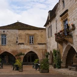 Bâtiments médiévaux en pierre avec arcades entourant une place à Monpazier. Balcon fleuri et tables en terrasse visibles.