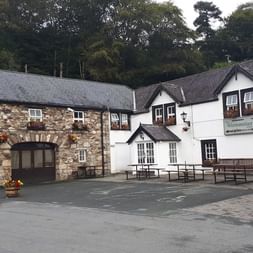 Glenmalure Lodge à Wicklow, Irlande : bâtiment blanc à deux étages avec annexe en pierre, bancs en bois dans la cour, entouré d'arbres.