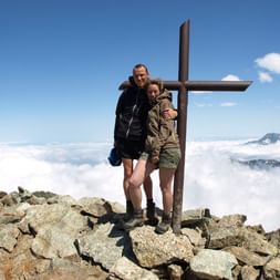 Deux randonneurs s'embrassent près d'une croix de sommet en bois sur un pic rocheux dans le Queyras, avec nuages et sommets visibles.