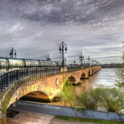 Pont de pierre avec lampadaires ornés enjambant la Garonne à Bordeaux. Un tramway moderne traverse le pont sous un ciel nuageux dramatique.