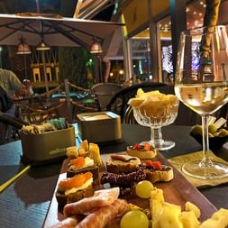 Planche en bois avec antipasti italiens : bruschetta, prosciutto, fromage, olives et raisins. Verre de vin blanc et chips sur terrasse.