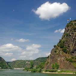 Falaise rocheuse escarpée avec drapeau allemand surplombant le Rhin près de Rüdesheim-Goarshausen. Collines boisées vertes et bâtiments blancs visibles.