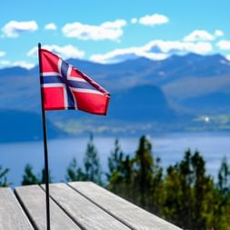 Drapeau norvégien sur terrasse en bois avec fjord et montagnes bleues en arrière-plan sous ciel clair. Végétation verte au premier plan.