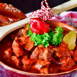 Bol de goulash hongrois garni de persil frais, paprika rouge et feuille de laurier, servi avec des bretzels.