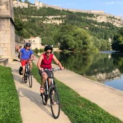 Cyclistes sur chemin pavé au bord d'une rivière calme en Franche-Comté. Falaises calcaires avec remparts et végétation verte dominent l'eau.