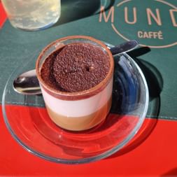 Tiramisu servi dans une tasse en verre sur soucoupe avec cuillère, montrant des couches distinctes de crème et cacao, sur un set Mundi Caffè.