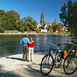 Cyclistes au bord du Danube à Ulm Deux cyclistes debout avec leurs vélos au bord du Danube à Ulm, admirant la cathédrale historique et la vieille ville sous un ciel bleu.