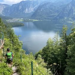 Randonneur avec sac à dos vert sur sentier de montagne surplombant un lac dans le Salzkammergut, Autriche, entouré de montagnes boisées.