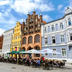 Bâtiments historiques colorés entourant une place de marché à Stralsund avec terrasses de café et visiteurs sous un ciel bleu nuageux.