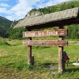 Panneau en bois 'Refuge des Fonts alt 2035' dans vallée alpine verte avec refuge traditionnel et pentes boisées du Queyras.