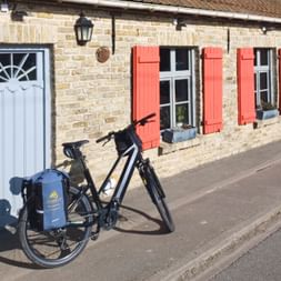 Vélo de randonnée noir avec sacoches bleues garé dans la rue à West-Cappel. Derrière, maison en briques avec volets corail et porte bleu pâle.