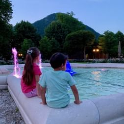 Deux enfants assis au bord d'une fontaine illuminée avec jets d'eau colorés à Bad Ischl. Montagnes boisées et ciel du soir en arrière-plan.
