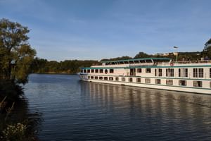 Bateau de croisière MS Switzerland naviguant sur un fleuve calme avec des arbres verts sur la rive gauche et des bâtiments visibles au loin.