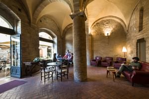 Lobby de l'Hôtel La Cisterna avec colonnes en pierre et plafonds voûtés, clients assis sur fauteuils en cuir et tables.