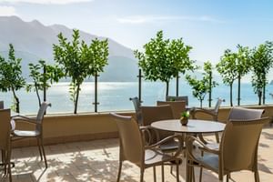 Terrasse extérieure de Villa Dirce Limone avec chaises beiges et tables rondes, petits arbres en pot, vue sur un lac avec montagnes en arrière-plan.