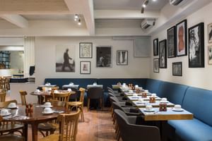 Intérieur de restaurant moderne avec banquette bleue, tables en bois dressées avec assiettes et tasses blanches, œuvres encadrées.