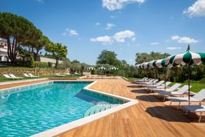 Piscine extérieure de la Villa Ermellina Siena avec terrasse en bois, chaises longues blanches, parasols rayés verts et paysage méditerranéen.