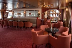 Salon élégant à bord du MS Switzerland avec fauteuils corail, tables rondes, tapis rouge et lustres décoratifs. Une arche mène à la salle à manger.