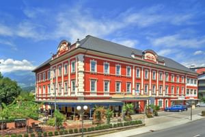 Hôtel Ertl Spittal, bâtiment rouge à trois étages avec garnitures blanches et toit gris. Terrasse au rez-de-chaussée avec sièges extérieurs sous ciel bleu.