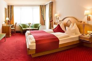 Chambre double élégante avec lit en bois, tapis rouge, fauteuils verts près de la fenêtre et décor alpin traditionnel à l'Hôtel Brückenwirt.