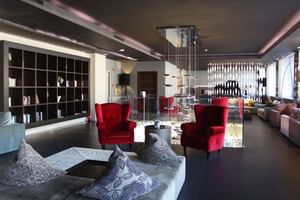 Hall d'entrée moderne C-Hotels avec sols sombres, fauteuils en velours rouge, tables en verre, étagères intégrées et éclairage contemporain.