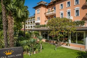 Élégant Hôtel Regina Adelaide de couleur corail avec balcons blancs, entouré de palmiers et jardins entretenus sous ciel bleu.