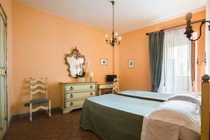 Chambre double de l'Hôtel La Cisterna avec murs orange, couvre-lit vert, miroir orné, commode antique, lustre et rideaux verts.