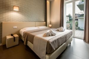 Chambre d'hôtel moderne au Palazzo del Garda avec lit double beige, mur texturé, cubes de chevet, applique murale et porte-fenêtre.