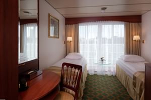 Cabine double sur le pont supérieur du MS Switzerland avec deux lits simples, table en verre, bureau en bois, miroir et grandes fenêtres avec rideaux beiges.