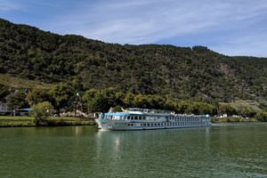 Bateau de croisière MS Switzerland naviguant sur eau verte calme avec collines boisées et petit village en arrière-plan sous ciel bleu.