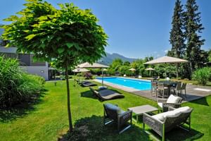Jardin de l'Hôtel Ertl à Spittal avec piscine bleue, chaises longues, mobilier d'extérieur et parasols sur pelouse verte avec montagnes en arrière-plan.