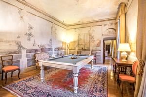 Salle de billard élégante avec table blanche sur tapis persan, murs ornés de fresques paysagères, chaises antiques et mobilier classique.