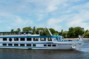 Bateau de croisière fluviale blanc MS Switzerland naviguant sur eau calme avec deux ponts de fenêtres, arbres verts et ciel bleu en arrière-plan.