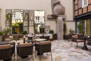 Cour d'hôtel moderne avec chaises en osier, tables rondes, sculpture en pierre et maisons à colombages traditionnelles à Colmar.