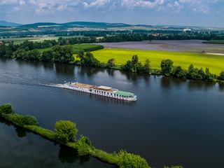 Vue aérienne du navire de croisière MS Florentina naviguant sur une large rivière entourée de champs verts, d'arbres et de collines sous un ciel nuageux.