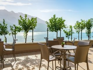 Terrasse extérieure de Villa Dirce Limone avec chaises beiges et tables rondes, petits arbres en pot, vue sur un lac avec montagnes en arrière-plan.