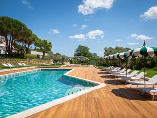 Piscine extérieure de la Villa Ermellina Siena avec terrasse en bois, chaises longues blanches, parasols rayés verts et paysage méditerranéen.