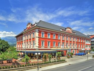 Hôtel Ertl Spittal, bâtiment rouge à trois étages avec garnitures blanches et toit gris. Terrasse au rez-de-chaussée avec sièges extérieurs sous ciel bleu.